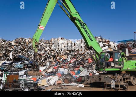 Kranlader mit Greiferarm und Magnet-Entladeanhänger vor dem Stapel von ausrangierten Autos, Haushaltsgeräten aus Metall und Industriegütern. Stockfoto