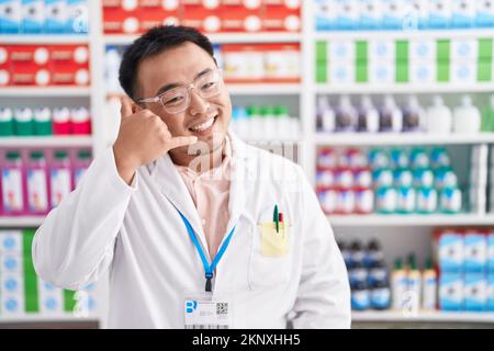 Chinesischer junger Mann, der in einer Apotheke arbeitet und lächelt und mit Hand und Fingern eine Telefongeste macht, als würde er am Telefon sprechen. Kommunikationskonzept Stockfoto