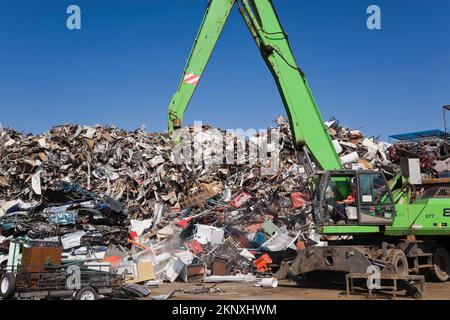 Kranlader mit Greiferarm und Magnet-Entladeanhänger vor dem Stapel von ausrangierten Autos, Haushaltsgeräten aus Metall und Industriegütern. Stockfoto