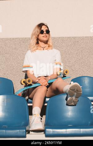 Stilvolle junge, legere Frau in Shorts mit Sonnenbrille und einem weißen T-Shirt mit Longboard sitzt im Sommer auf den Stadion-Tribünen der Stadt Stockfoto