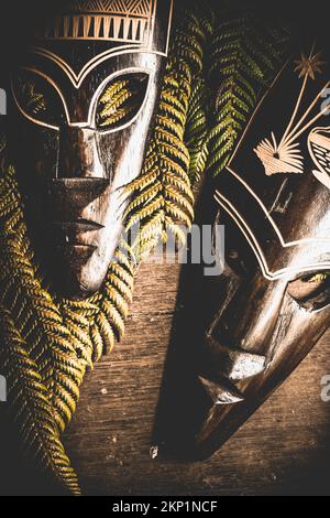Foto des kulturellen Stilllebens auf afrikanischen traditionellen Stammesmasken mit Regenwaldfarnen auf altem Holztisch. Kampf der Zivilisationen Stockfoto