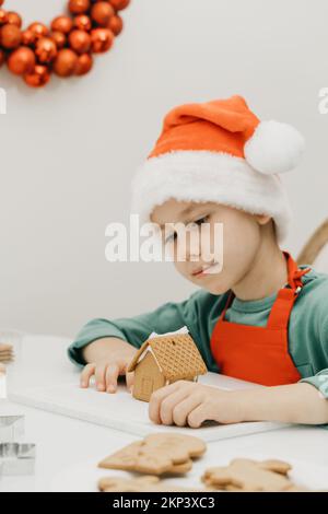 Ein süßer Junge an Silvester und Weihnachten macht in einem Weihnachtshut ein Ingwerhaus in der Küche. Stockfoto