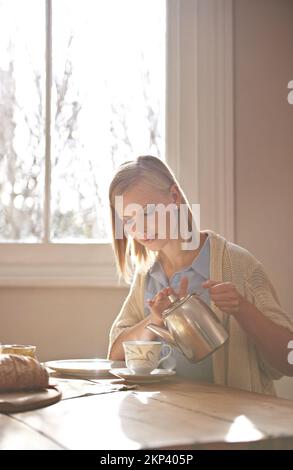 Unterschätze niemals die Kraft einer guten Tasse Tee. Eine schöne junge Frau, die sich eine Tasse Tee einschenkt, während sie zu Hause am Tisch sitzt. Stockfoto
