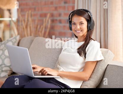 Mit ihren Online-Freunden in Verbindung treten. Eine attraktive junge Frau, die Kopfhörer trägt, während sie ihr Notebook zu Hause benutzt. Stockfoto