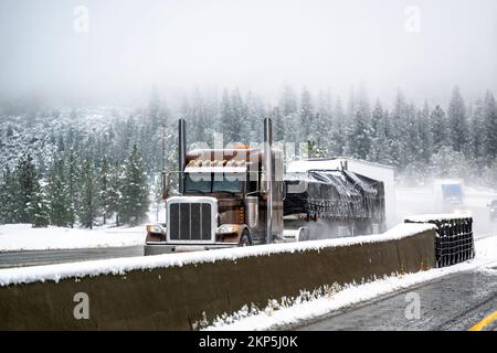 Klassischer brauner Sattelschlepper mit eingeschalteten Scheinwerfern transportiert abgedeckte kommerzielle Fracht auf einem Tieflader-Sattelanhänger, der vor Anothe fährt Stockfoto