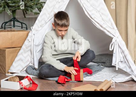 Geschenkverpackung. Ein Teenager verpackt Weihnachtsgeschenke in Kraftpapier mit einem Seil und roten Bändern. Ein ernster Junge packt Geschenke für die Familie. Fa Stockfoto