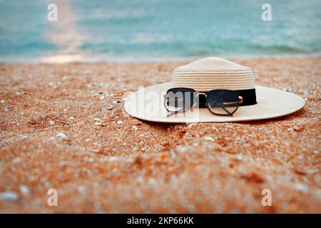 Am Strand liegt ein Strohhut mit schwarzer Sonnenbrille. Familienurlaub im Hotel. Entspannen Sie sich am Strand. Werbung in Reisebüros. Hochwertiges Foto. Stockfoto
