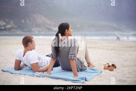 Die Landschaft mit diesem besonderen Menschen zu genießen. Rückblick auf ein junges Paar, das am Strand sitzt. Stockfoto