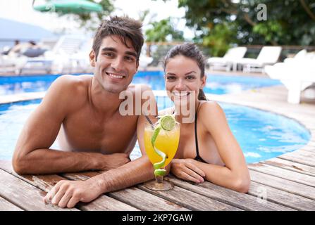 Einen Drink teilen. Ein attraktives junges Paar ruht sich am Rand eines Pools mit einem Cocktail aus. Stockfoto