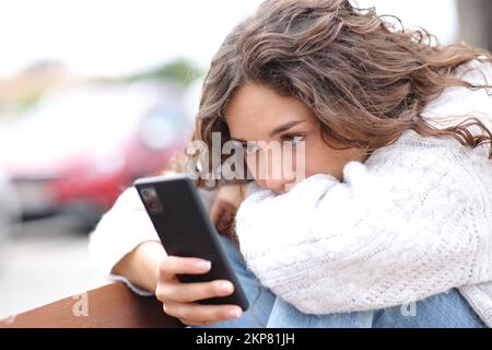 Eine traurige Frau, die auf dem Smartphone sitzt und auf einer Bank auf der Straße sitzt Stockfoto