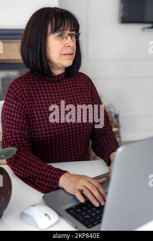 Attraktive Frau mittleren Alters, die an einem Laptop arbeitet, während sie an einem Tisch sitzt. Stockfoto