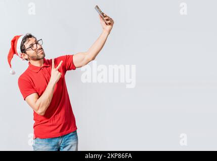 Lächelnder junger Mann mit weihnachtsmütze, der ein Selfie in Einzelhaft macht. Ein glücklicher gutaussehender Mann mit weihnachtsmütze, der ein Selfie macht. Ein glücklicher Kerl, der ein weihnachts-Selfie macht Stockfoto
