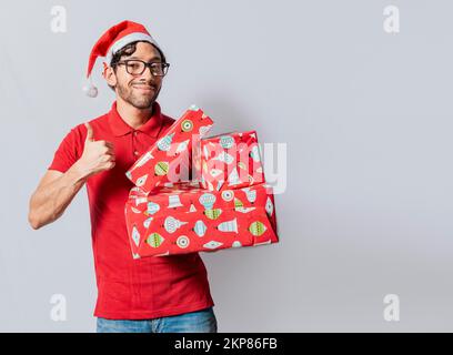 Weihnachtsmann mit Geschenkschachteln und lächelndem Daumen nach oben. Ein lächelnder Typ mit weihnachtsmütze, der Weihnachtsgeschenkschachteln hält und Daumen nach oben zeigt. Freundlicher Mann i Stockfoto