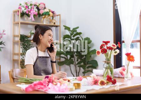 Floristen-Konzept, weibliche Floristin im Gespräch mit dem Kunden am Telefon und Notizen im Notebook im Geschäft. Stockfoto