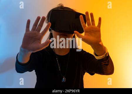 Ein Mädchen im Teenageralter mit Haaren, die eine Virtual-Reality-Brille tragen, schaut sich Filme an oder spielt Videospiele, zeigt ein Zeichen der Dankbarkeit mit ihren Händen. Lustiges Mädchen Stockfoto