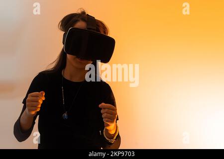 Ein Mädchen im Teenageralter mit Haaren, die eine Virtual-Reality-Brille tragen, schaut sich Filme an oder spielt Videospiele, zeigt ein Zeichen der Dankbarkeit mit ihren Händen. Lustiges Mädchen Stockfoto