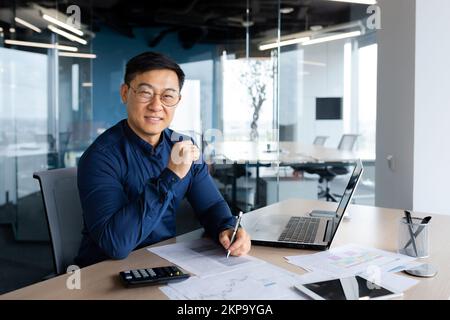 Portrait erfolgreicher, zufriedener asiatischer Finanzbuchhalter, Mann im Hemd und mit Brille, der im Büro mit Taschenrechner und Laptop für Berichte über Konten und Finanzdokumente arbeitet, und auf die Kamera schaut. Stockfoto