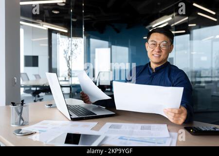Portrait erfolgreicher, zufriedener asiatischer Finanzbuchhalter, Mann im Hemd und mit Brille, der im Büro mit Taschenrechner und Laptop für Berichte über Konten und Finanzdokumente arbeitet, und auf die Kamera schaut. Stockfoto