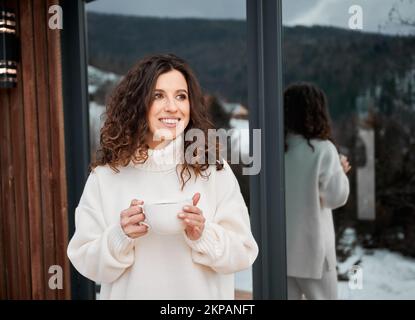 Lockige junge Frau, die auf der Terrasse des modernen Scheunenhauses in den Bergen ruht. Glückliche weibliche Tourist mit einer Tasse Tee, genießen in neuen Hütte im Winter. Stockfoto