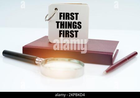 Geschäfts- und Finanzkonzept. Auf einer weißen Oberfläche liegen ein rotes Notizbuch und ein Stift, eine Lupe und ein weißes Notizbuch mit der Aufschrift „First Thi“ Stockfoto