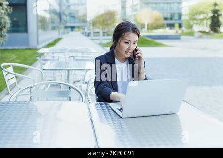 Asiatische selbstbewusste Geschäftsfrau, die telefoniert und im Freien mit einem Laptop arbeitet. Job, Job Aspiration Konzept. Geschäftsleute, Lebensstil, Kopierraum Stockfoto