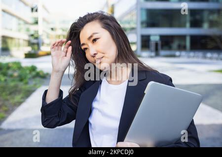 Müde, überarbeitete asiatische Geschäftsfrau im Anzug mit Laptop. Stress am Arbeitsplatz, Migräne, Frustration und Depression Konzept, Hintergrund des Bürozentrums. Geschäftsleute, Lebensstil, Kopierraum Stockfoto