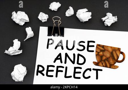 Ruhe bei der Arbeit. Auf der schwarzen Oberfläche befindet sich ein weißes Blatt mit der Aufschrift – PAUSE UND REFLEXION – eine Tasse mit einem Bild von Kaffeebohnen. Stockfoto