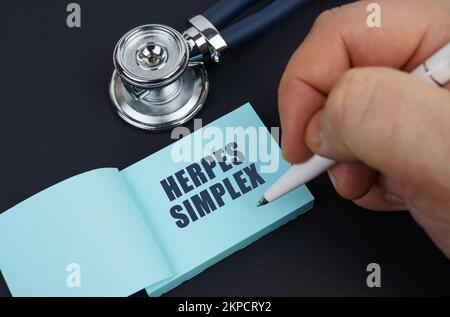 Medizin und Gesundheitskonzept. Auf einem schwarzen Tisch sind ein Stethoskop und ein Notizbuch, in dem der Mann geschrieben hat - HERPES SIMPLEX Stockfoto