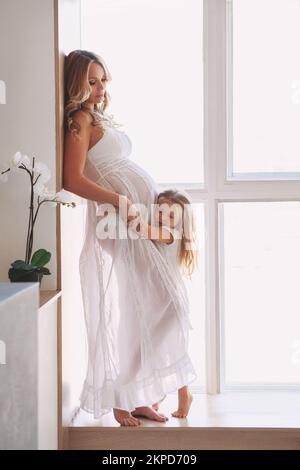 Wunderschöne schwangere mutter mit Kind, das zu Hause am Fenster posiert. Ein Mädchen, das den Bauch umarmt und dem Baby zuhört. Modemodelle tragen lange weiße Kleider Stockfoto
