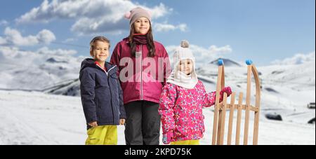 Kinder in Winterkleidung stehen mit einem hölzernen Schlitten auf einem verschneiten Berg Stockfoto