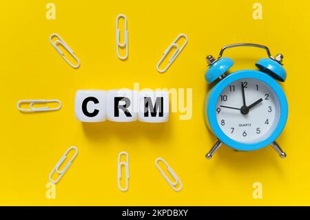 Finanz- und Wirtschaftskonzept. Auf gelbem Hintergrund, blauer Wecker, Büroklammern und weiße Würfel, auf die der Text geschrieben ist – CRM Stockfoto