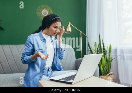 Seriöse junge hispanische Frau, die von zu Hause aus arbeitet, Beratung, Helpdesk, Kundenservice. Sitzen auf dem Sofa mit einem Laptop und Kopfhörern während eines Videogesprächs. Stockfoto