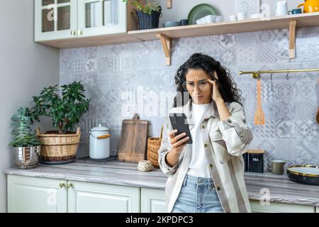 Schockierte Lateinamerikanerin steht zu Hause in der Küche und hält das Telefon in der Hand. Verärgert, schaut sie auf ihr Handy, hat unerwartete und schlechte Nachrichten erhalten, Nachrichten. Stockfoto