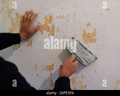 Abkratzen von Tapetenresten von der Wand mit einem Pfannenwender durch einen Mann in schwarzer Kleidung, Vorbereitung und Reinigung der Wandoberfläche vor dem Tapeten Stockfoto