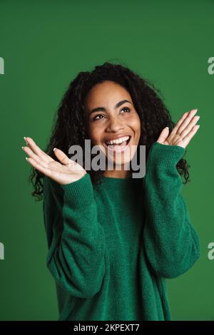 Junge schwarze Frau, die einen Pullover trägt, lacht und Gesten, isoliert vor grünem Hintergrund Stockfoto