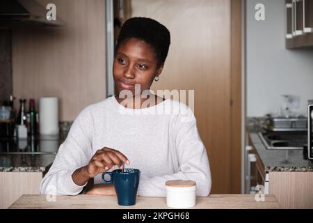 Träumige afroamerikanische Frau, die in der Küche sitzt und Tee rührt Stockfoto