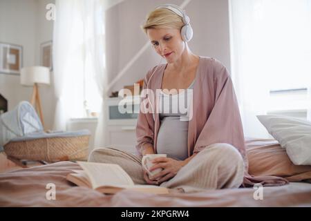 Schwangere Frau, die in ihrem Bett sitzt, Musik hört, Bücher liest und sich selbst Zeit nimmt. Stockfoto