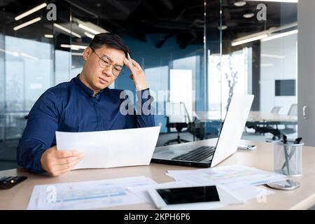 Ein verzweifelter Geschäftsmann sieht und liest Finanzberichte und Dokumente, der asiatische Chef arbeitet mit Dokumenten im Büro, verwirrt und deprimiert. Stockfoto