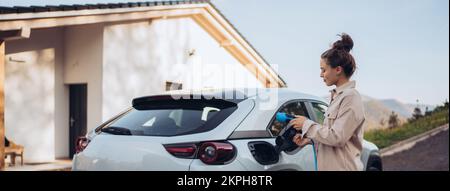 Junge Frau, die das Stromkabel von ihrem Elektroauto hält, bereit, es zu Hause zu laden, ein nachhaltiges und wirtschaftliches Transportkonzept. Stockfoto