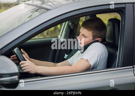 Ein junger Kerl fährt unsicher, während er vor dem Lenkrad telefoniert. Der Fahrer verwendet sein Smartphone. Risiko- und Gefahrensituationen auf dem Roa Stockfoto