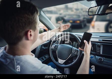 Wütender und unzufriedener Fahrer, der mit der Hand den Stau vor seinem Auto sieht. Aggressiver und unzufriedener Kerl, unsicheres Fahren, da er sein Handy i benutzt Stockfoto