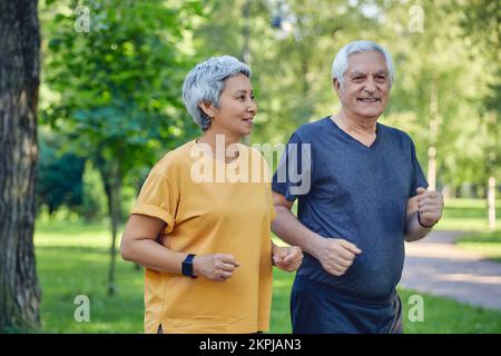 Reifes, grauhaariges Paar, das im Sommerpark joggt. Zwei ältere Jogger genießen morgens aktive Kardio-Aktivitäten im Freien, halten einen gesunden aktiven Lebensstil. Wiegen Stockfoto