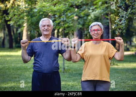 Ältere 60 Ehepartner tragen Sportbekleidung, die sich mit Handübungen auseinandersetzt, und trainieren morgens im Sommerpark mit Gummibändern im Freien. Gesundes li Stockfoto