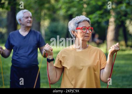 Ältere 60 Ehepartner tragen Sportbekleidung, die sich mit Handübungen auseinandersetzt, und trainieren morgens im Sommerpark mit Gummibändern im Freien. Gesundes li Stockfoto