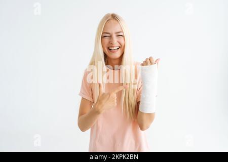 Lachende blonde junge Frau, die Zeigefinger auf einen gebrochenen Arm zeigt, der in Pflasterbandage gewickelt ist, in die Kamera schaut, auf einem weißen, isolierten Hintergrund steht. Stockfoto