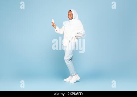 Glückliche afroamerikanische Frau in Hidschab, die Handy benutzt, auf blauem Studiohintergrund steht, freier Platz, volle Körperlänge Stockfoto