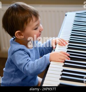 Kleinkind Baby spielt Klavier im Wohnzimmer, Musikunterricht. Glücklicher Junge lernt, Musik auf einem elektrischen Klavier zu spielen. Kind Junge im Alter von einem Jahr vier mont Stockfoto