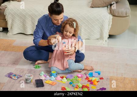 Mutter hilft der kleinen Tochter, Seifenblasen zu machen, wenn sie im Kinderzimmer auf dem Boden sitzt Stockfoto