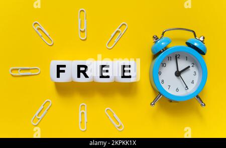 Finanz- und Wirtschaftskonzept. Auf gelbem Hintergrund, blauer Wecker, Büroklammern und weiße Würfel, auf denen der Text geschrieben ist – KOSTENLOS Stockfoto