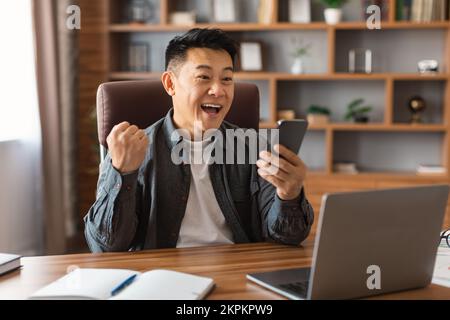 Glücklicher japanischer Geschäftsmann mittleren Alters mit offenem Mund, der auf sein Smartphone schaut und eine Siegesgeste macht Stockfoto